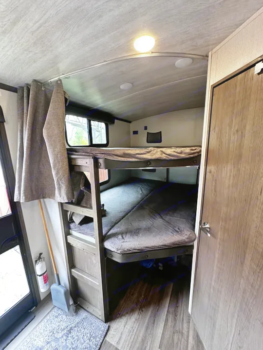 Cozy RV rental bunk beds, privacy curtains pulled aside. Features a fire extinguisher, broom, and ceiling light for comfy travel.. Keystone RV Cougar 2023