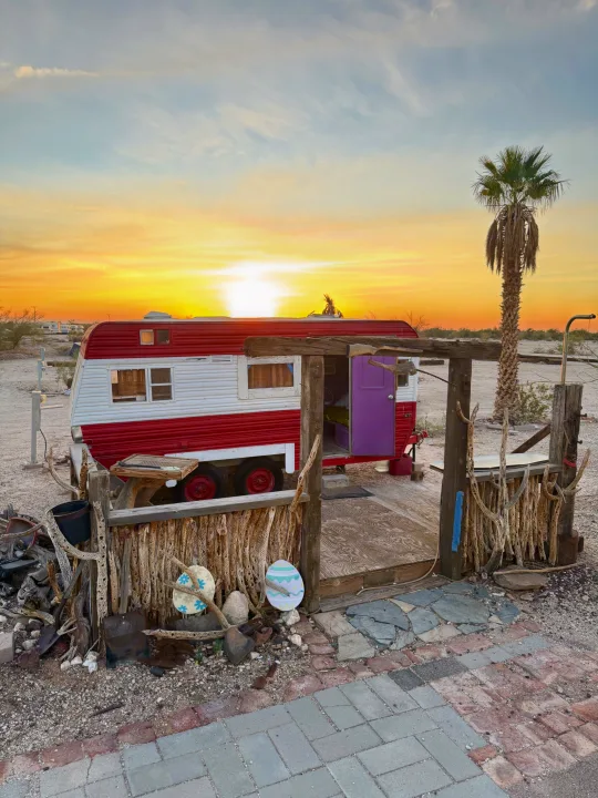 Vintage red & white rv rental set in the desert. Features a unique wooden porch, purple door, and a vibrant sunset.. Kit Kit Trailer Deluxe 1978