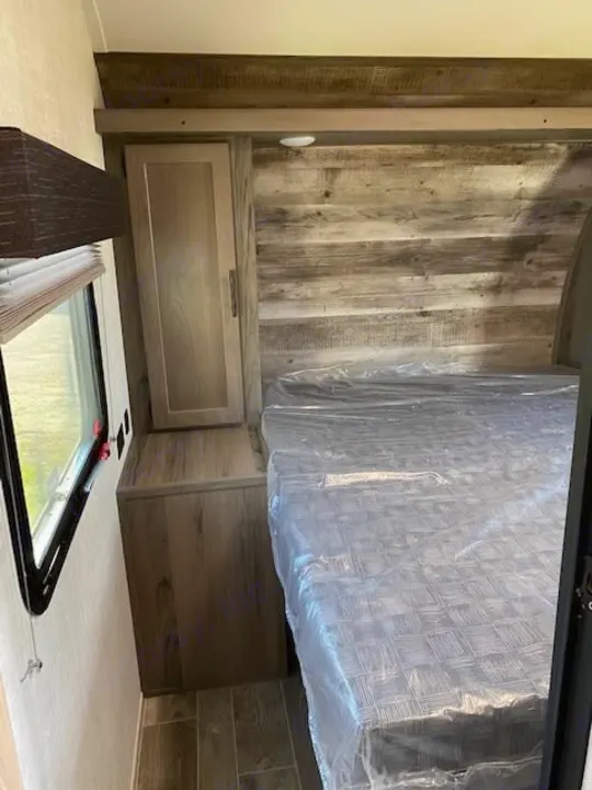 RV rental bedroom shows a bed with plastic, window, cabinet, and rustic wood paneling. Ready for guests.. Gulf Stream Ameri-Lite 2023
