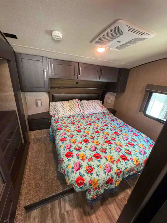 Cozy RV rental bedroom featuring a vibrant floral bedspread, overhead cabinets, window, and ceiling AC unit for comfort.. Grand Design Reflection 2022