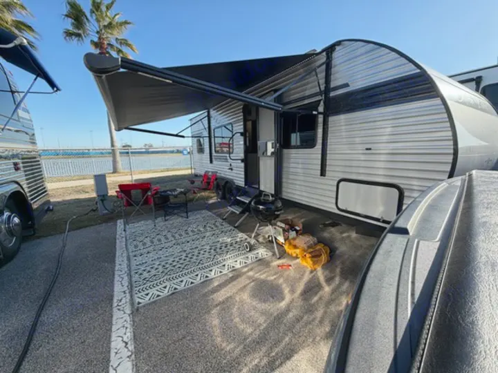 RV rental campsite: a travel trailer with an extended awning, outdoor rug, grill, and chairs, overlooking a sunny bay.. East to West Silver Lake 2024