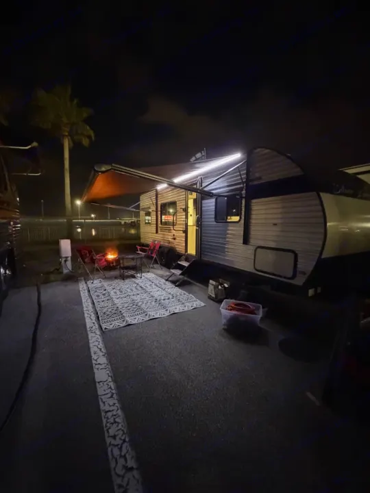 Cozy night at an RV rental campsite, featuring an RV with awning lights, outdoor rug, chairs, and a glowing fire pit.. East to West Silver Lake 2024