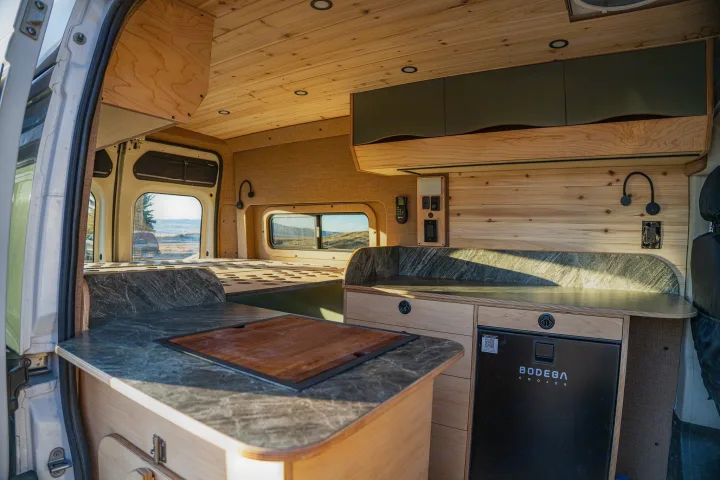 Bright, modern rv rental camper van interior with custom wood, kitchen counter, fridge, and bed area visible for travel.. Ram Promaster 2015