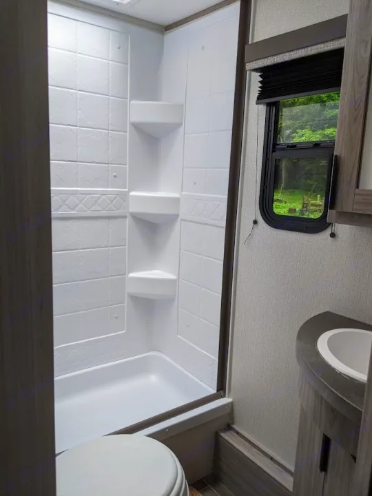 Clean RV rental bathroom features a compact white shower with shelves, toilet, and small vanity with sink next to a window.. Keystone RV Passport SL 2021