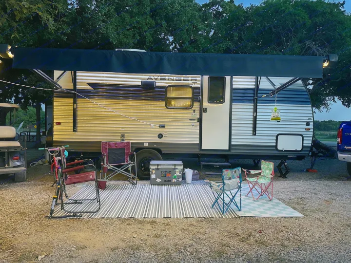 Cozy rv rental camping setup at dusk, featuring a travel trailer with extended awning, string lights, chairs, and outdoor rugs.. Prime Time Avenger - 17BHS 2022