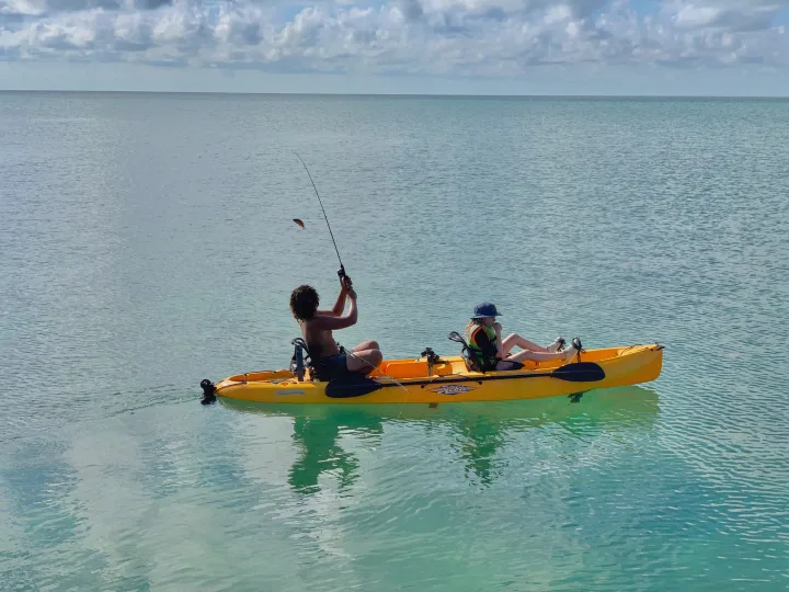 Two people kayak fishing, one reeling in a small fish on clear waters. A perfect water activity for your RV rental adventure!. Heartland RVs Pioneer 2022
