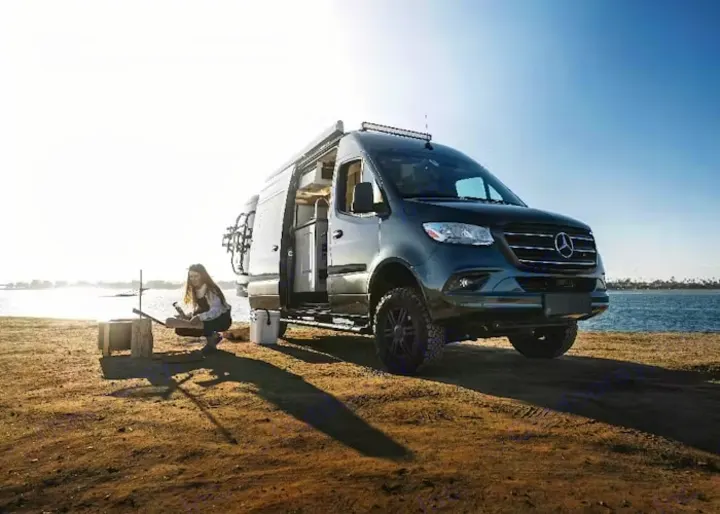 Explore nature with this stylish rv rental camper van by the water. A woman is preparing outdoors, embracing the van life adventure.. Thor Motor Coach Sprinter 4X4 Tranquility 2023