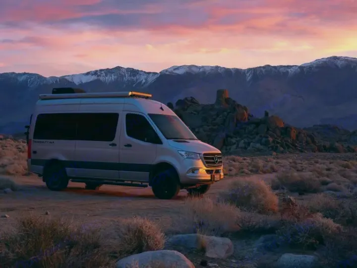 A silver Mercedes Sprinter rv rental, a Tranquility 4x4, rests in a desert landscape with snow-capped mountains at sunset.. Thor Motor Coach Sprinter 4X4 Tranquility 2023