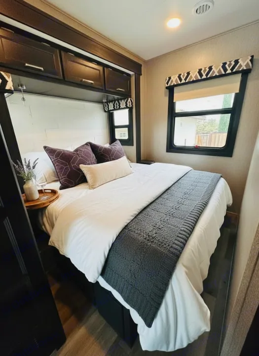 Cozy RV rental bedroom with white linens, purple pillows, gray throw, overhead cabinets, and windows for natural light.. Jayco Alante 2021