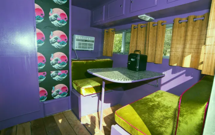 Vibrant purple and olive green interior of an RV rental, featuring retro wave wallpaper, a dining nook, and a mini-fridge.. Kit Kit Trailer Deluxe 1978