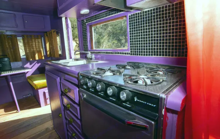Vibrantly purple RV rental kitchen featuring a stove, sink, checkered backsplash, and a view through the window.. Kit Kit Trailer Deluxe 1978