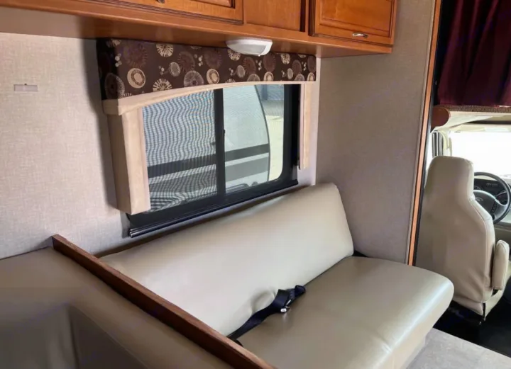 This RV rental interior features a light-colored dinette bench, window with shade, overhead cabinets, and view of the driver's seat.. Winnebago Minnie Winnie 2015