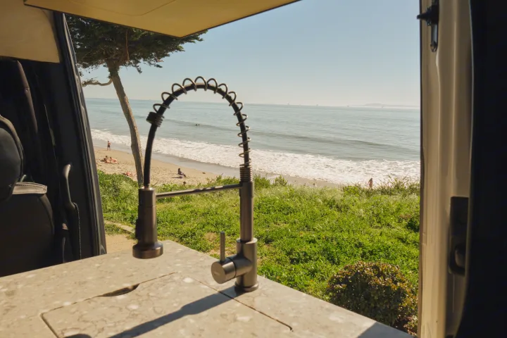 RV rental with a seaside kitchen view: modern faucet, beach with sunbathers, crashing waves, distant ocean, and offshore platforms.. Mercedes Sprinter 2022