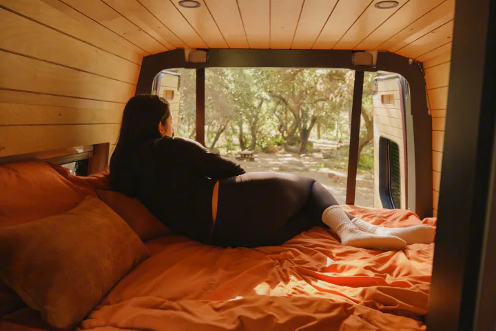 A woman relaxes in a cozy rv rental, gazing from orange bedding out to sunlit trees from the custom-built van.. Mercedes Sprinter 2022