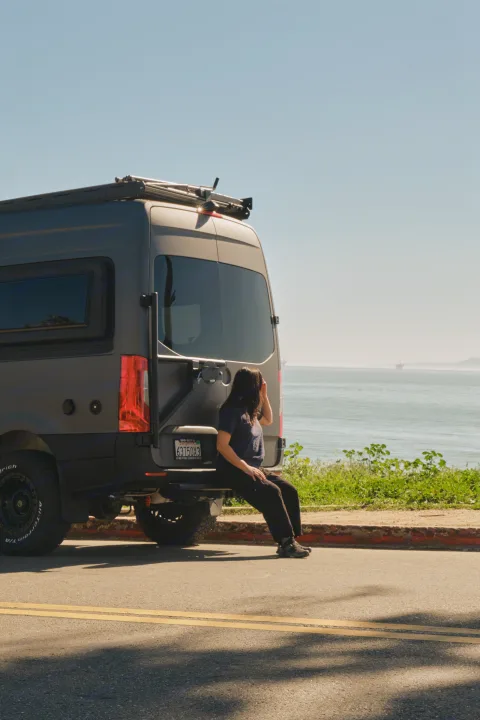A person sits on a dark gray camper van's bumper, facing the ocean. An ideal rv rental for a scenic road trip.. Mercedes Sprinter 2022
