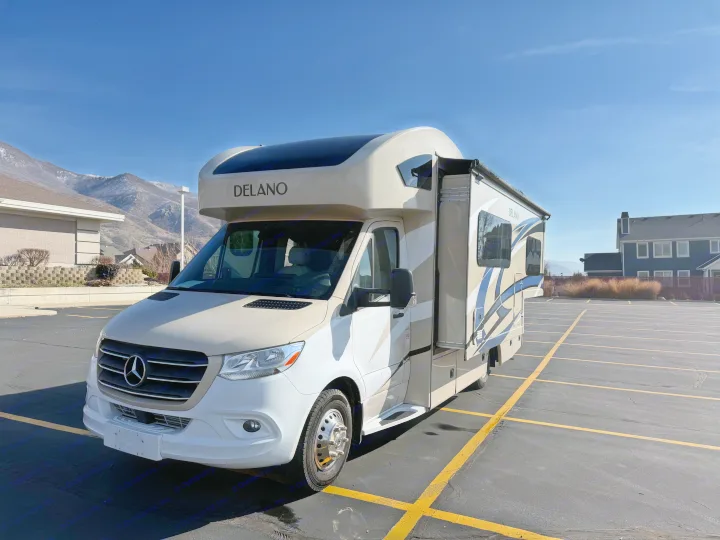 A beige and white Delano Mercedes-Benz RV rental sits in a sunny parking lot before mountains.. Thor Delano 2021