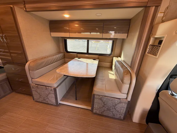 RV rental interior: A light-toned U-shaped dinette with a table, overhead cabinets, and a large window offers comfortable seating.. Thor Delano 2021
