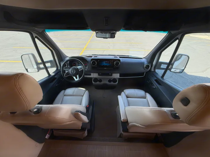 RV rental cockpit: light leather driver & passenger seats, Mercedes-Benz dashboard, and a view of a parking lot.. Thor Delano 2021