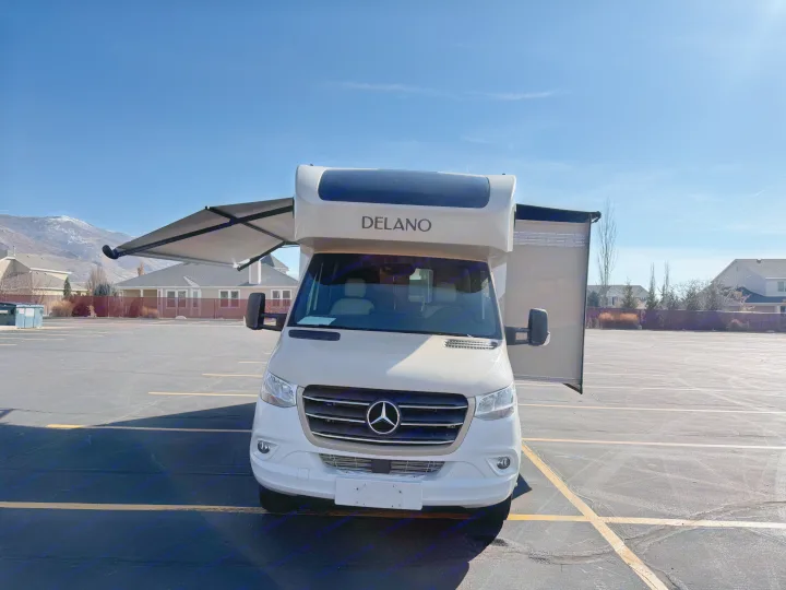 This beige Mercedes-Benz Delano RV rental is parked with its awning extended in a lot, with mountains in the background.. Thor Delano 2021