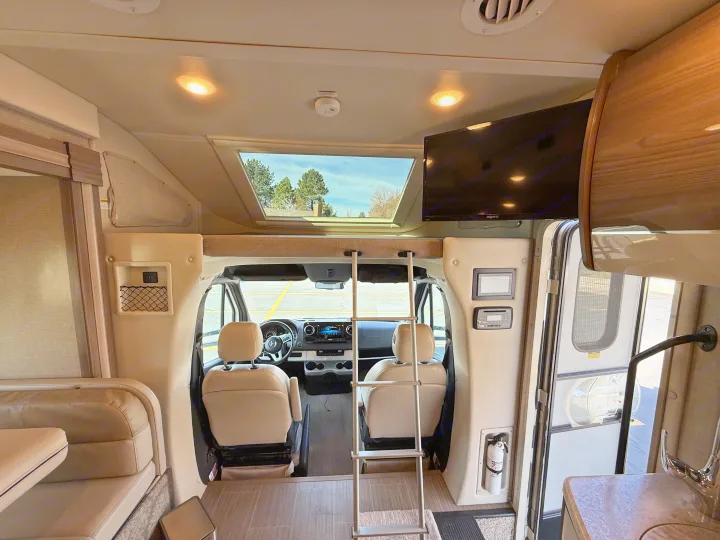 Spacious RV rental interior showing the cockpit, overhead bunk with ladder, and living area with a TV.. Thor Delano 2021
