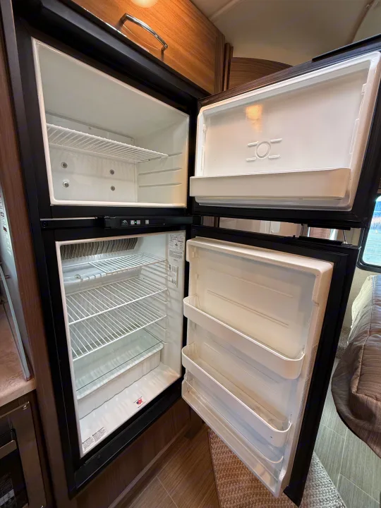 Clean, empty two-door refrigerator/freezer inside an RV rental, showing shelves and door bins, ready for your trip.. Thor Delano 2021