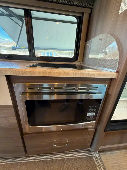 Compact kitchen in an RV rental, featuring a countertop, built-in cooktop, microwave, and window with an exterior awning.. Thor Delano 2021