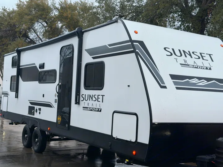 A clean, white Sunset Trail Sport travel trailer, perfect for rv rental, stands wet outdoors, featuring black trim and dual axles.. Crossroads RV Sunset Trail Sport 2025