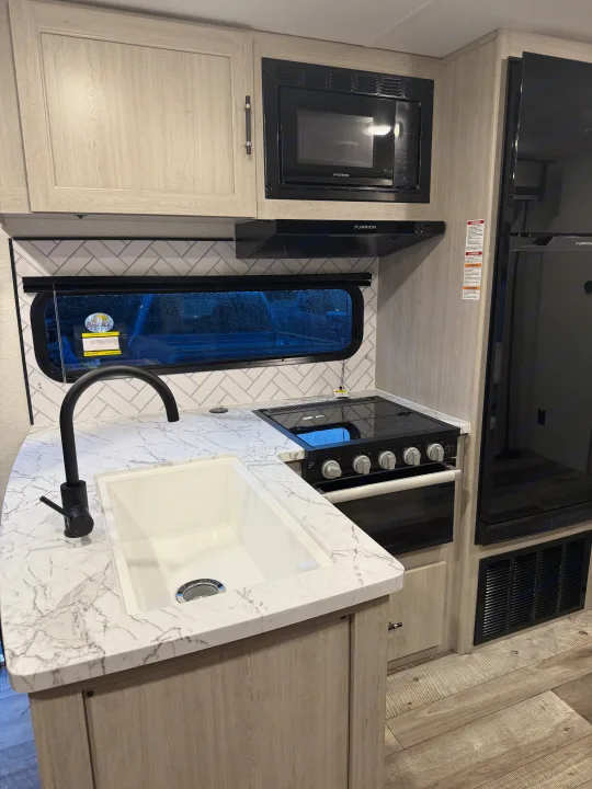 A sleek RV rental kitchen features a white marbled countertop, black faucet, stove, microwave, and light wood cabinetry with tile backsplash.. Crossroads RV Sunset Trail Sport 2025