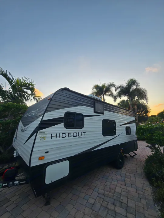 This Hideout by Keystone travel trailer, ideal for an RV rental, is parked on pavers at sunset, surrounded by lush palm trees.. Keystone RV Hideout 2021