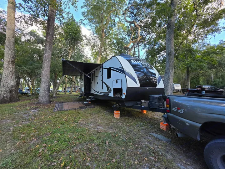White RV rental trailer with awning extended, hitched to a pickup truck in a wooded campground. Orange leveling blocks visible.. Crossroads RV Sunset Trail Super Lite 2018