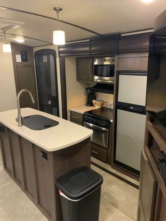 Modern RV rental kitchen features an island, sink, stove, microwave, and fridge. Compact, dark cabinetry, and white counters.. Crossroads RV Sunset Trail Super Lite 2018