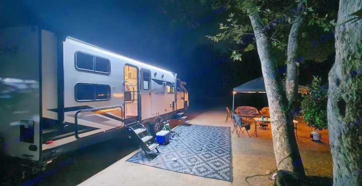 Evening camping: A bright RV rental illuminates a cozy outdoor living space with mat and chairs under dark trees.. Grand Design Imagine 2022