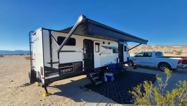 An Imagine RV rental is set up for desert camping with its awning extended, chairs, and truck under a clear blue sky.. Grand Design Imagine 2022