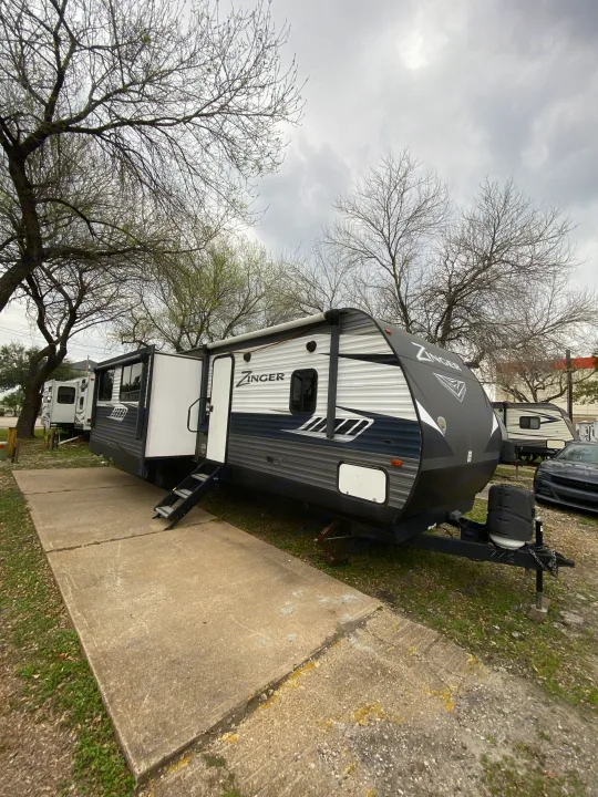 Zinger travel trailer RV rental set up at a park, with steps down & slide-out extended, under bare trees on an overcast day.. Crossroads RV Zinger 2019