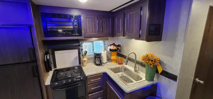 This modern RV rental kitchen showcases dark wood cabinets, a microwave, stove, sink, and blue accent lighting, ideal for travel.. Crossroads RV Zinger 2019