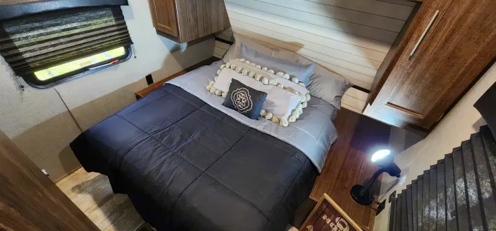 Cozy RV rental bedroom featuring a dark bed, decorative pillows, window with blind, and a functional reading lamp.. Crossroads RV Zinger 2019