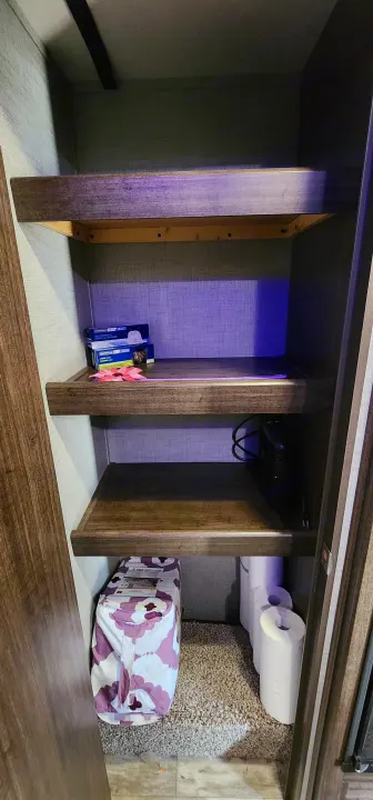 RV rental pantry: Three wooden shelves with kitchen bags, electronics, a patterned bag, and toilet paper rolls.. Crossroads RV Zinger 2019