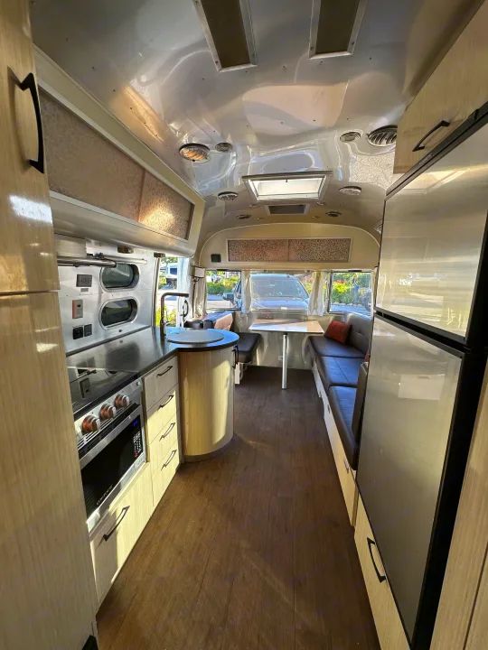 Spacious RV rental interior features modern kitchen, dining booth, fridge, dark wood flooring, and polished aluminum ceiling.. Airstream International 2020