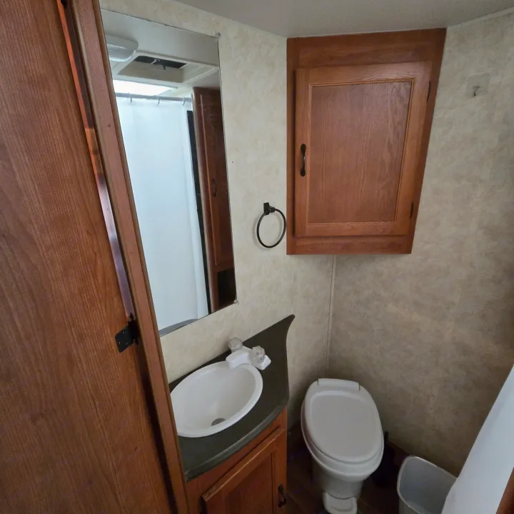 View of a functional RV rental bathroom with a white sink, small mirror, toilet, and wooden overhead cabinet.. Coachmen Freelander 2011