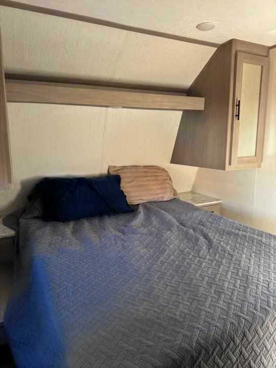 Inside an RV rental: a compact bedroom with a bed, blue and beige pillows, and gray quilted comforter, plus overhead storage.. Coleman Lantern 2022