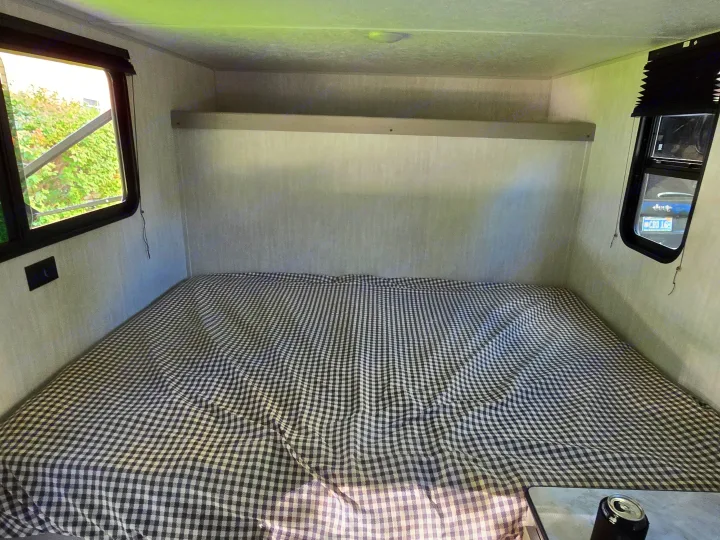 Inside an RV rental: a compact sleeping area with a checkered bed, overhead shelf, and windows showing nature & a Jeep.. Clipper 3K 2025