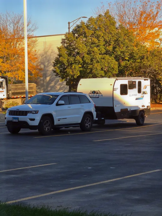 White Jeep Grand Cherokee towing a Clipper RV rental in a sunny parking lot, surrounded by autumn trees and a building.. Clipper 3K 2025