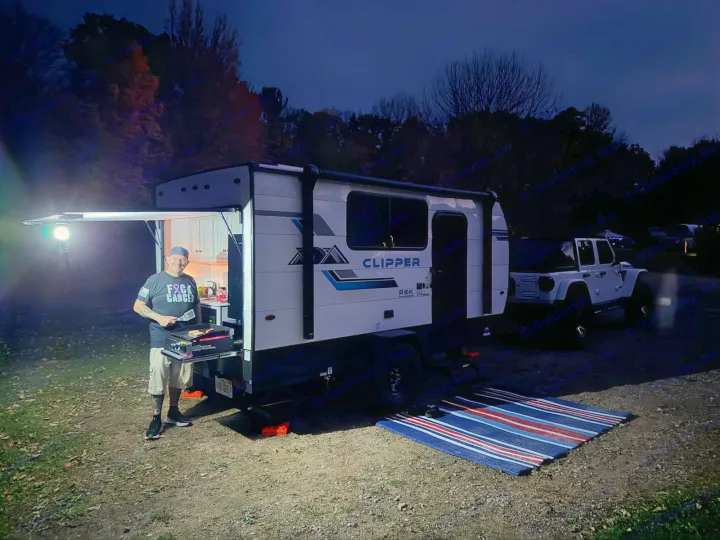 Enjoying an RV rental experience: a man cooks dinner outside a Clipper RV at a dark campsite, with a Jeep nearby.. Clipper 3K 2025