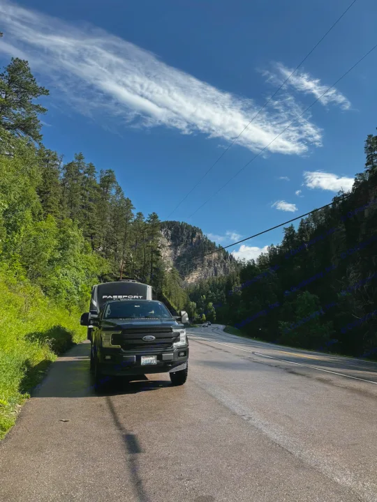 Black Ford truck towing an RV rental on a winding mountain road. Lush green trees, a rocky cliff, & blue sky enhance this scenic drive.. Keystone RV Passport Grand Touring 2019