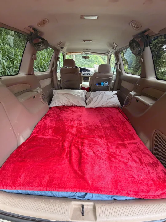 Minivan converted into an RV rental sleeping setup. Features a red blanket, pillows & fans for comfortable car camping.. Toyota Sienna 2002