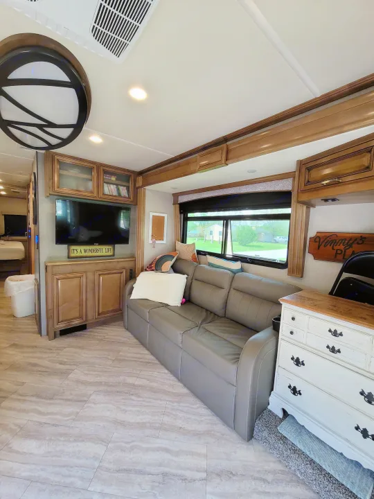 Interior of an RV rental with a gray sofa facing a TV, wood cabinets, and a window showcasing a neighborhood view.. Fleetwood Southwind 36P 2019