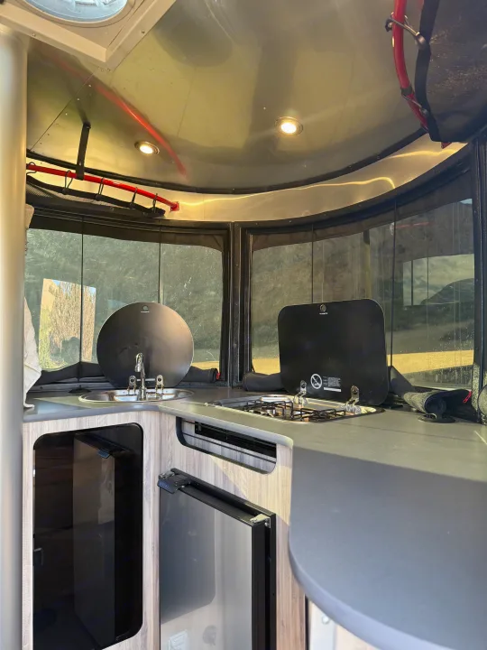 Inside a modern rv rental: compact galley featuring a covered sink, two-burner stove, and under-counter fridge.. Airstream Basecamp 2020