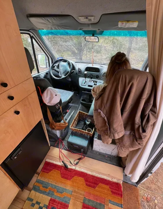RV rental camper van: wood cabinets, mini-fridge, power station, colorful rug. Person seated with trees visible outside.. Dodge Sprinter Van 2006