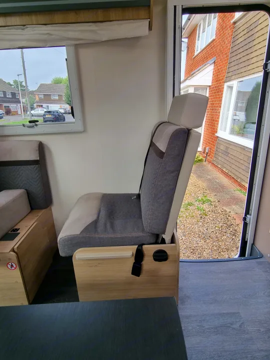 Step into this RV rental: a cozy grey passenger seat with seatbelt, light wood cabinetry, and a window view of a quiet street.. Chausson 720 VIP Titanium 2021