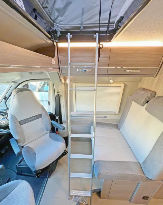 Inside an RV rental: swiveling driver's seat, ladder to pop-top bed, overhead cabinets, and dinette seating.. Elddis CV80 2024